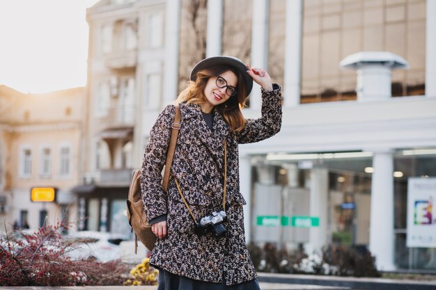 Открытый портрет элегантной молодой леди с коричневым рюкзаком носить пальто и шляпу. Привлекательная женщина с вьющимися волосами разговаривает по телефону, попивая кофе на улице