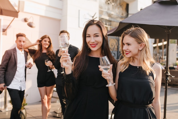 Outdoor portrait of enthusiastic brunette women in black dresses posing