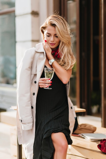 Открытый портрет модной женской модели в плиссированном платье пьет шампанское и смотрит вниз. Радостная блондинка в бежевом плаще держит бокал вина, стоя на улице в холодный день.