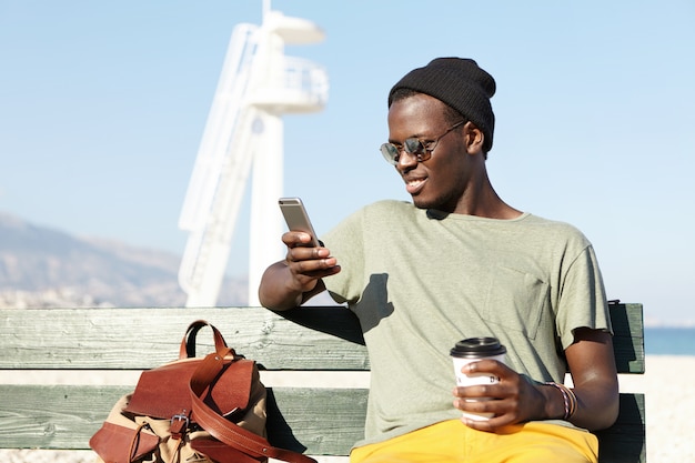 Ritratto all'aperto del turista maschio afroamericano sorridente alla moda che gode del caffè dalla tazza di carta e che cerca nuovi posti interessanti sui siti web facendo uso dell'applicazione online sul telefono cellulare