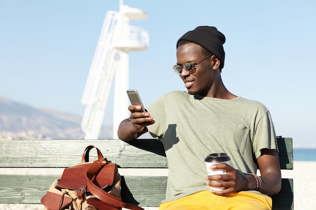 Outdoor portrait of fashionable smiling African American male tourist enjoying coffee out of paper cup and searching for new interesting places on websites using online application on mobile phone