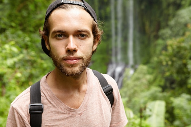 Outdoor portrait of fashionable young bearded man wearing black snapback backwards standing against exotic green nature with waterfall. Caucasian tourist spending vacations in rainforest