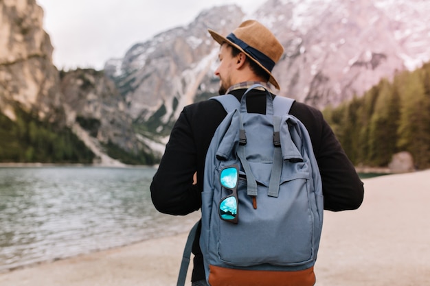 Outdoor portrait from back of male tourist carrying big decorated backpack and walking to mountains in morning