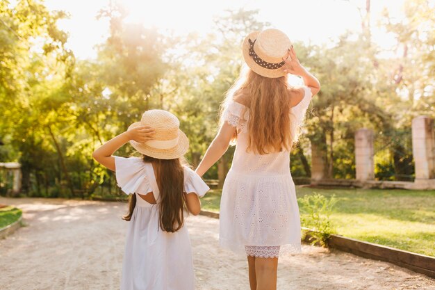 Outdoor portrait from back of tanned woman in white dress and her daughter in similar outfit enjoying lovely nature views. Pretty long-haired lady holding hands with little girl posing in sunlight.