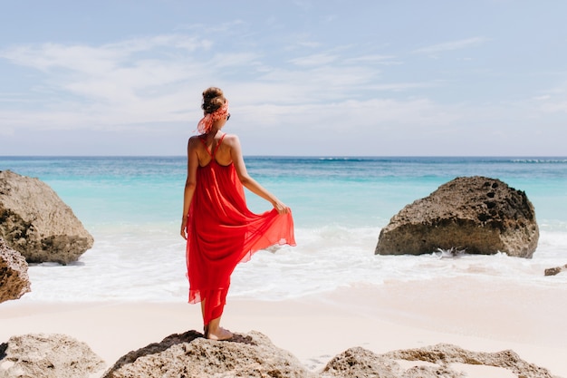 Outdoor portrait from back of wonderful caucasian woman enjoying sea views in weekend. Full-length shot of pleased girl in long dress looking at sky near ocean.