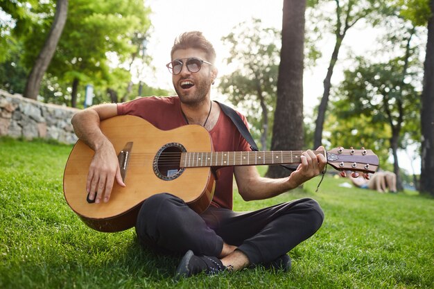 Outdoor portrait of handsome carefree hipster guy sitting on grass in park and playing guitar