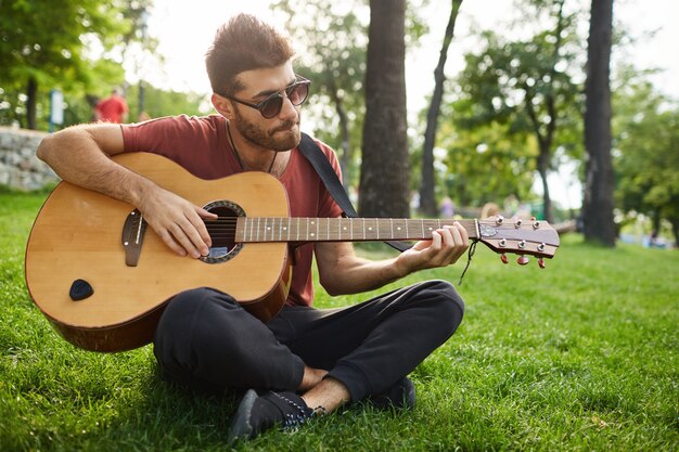 公園の芝生の上に座って、ギターを弾くハンサムな流行に敏感な男の屋外のポートレート