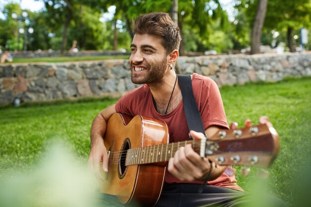 Outdoor portrait of handsome man smiling, sitting on grass in park and playing guitar