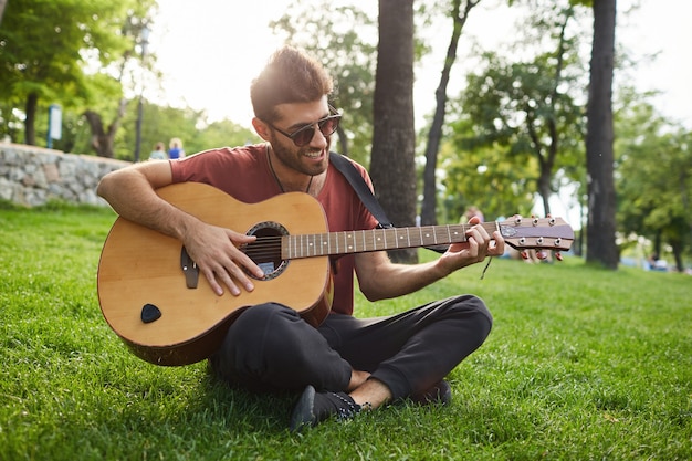 Outdoor portrait of handsome smiling hipster guy sitting on grass in park and playing guitar