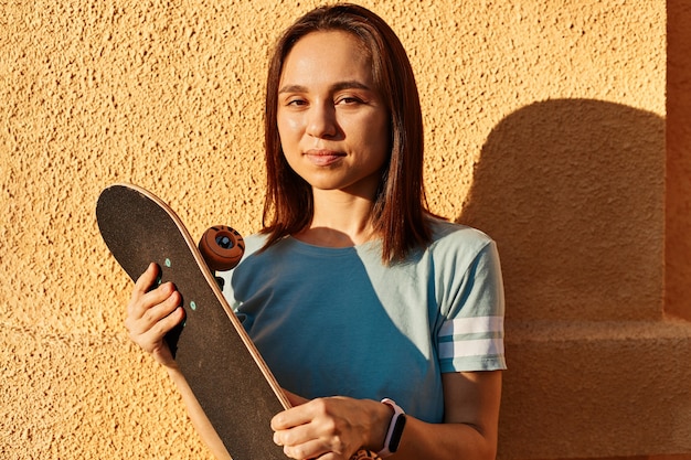 Outdoor portrait of happy dark haired female wearing t shirt posing isolated over yellow wall, looking art camera, holding skateboard in hands, summertime, sunset.