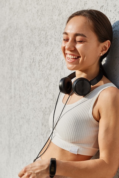 Free photo outdoor portrait of happy laughing female wearing white sports top, standing with headphones over neck