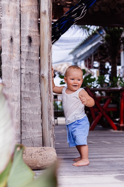 Outdoor portrait of happy nine months old kid in blue short and white shirt stands by wooden wall and smile