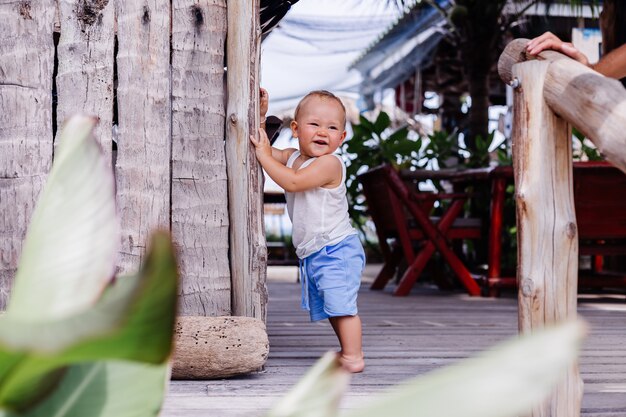 Outdoor portrait of happy nine months old kid in blue short and white shirt stands by wooden wall and smile