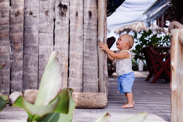 Outdoor portrait of happy nine months old kid in blue short and white shirt stands by wooden wall and smile