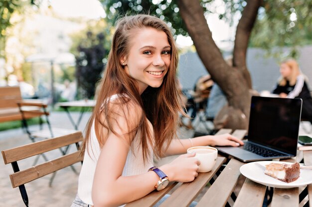 Открытый портрет счастливой улыбающейся девушки с длинными волосами и большими глазами, работающей на улице с ноутбуком