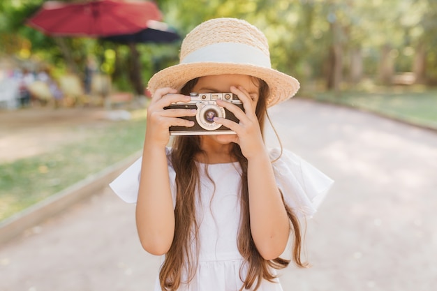 公園で時間を過ごし、自然の景色の写真を作るインスピレーションを得た少女の屋外の肖像画。道路に立っているカメラを保持している長い茶色の髪の帽子の子供