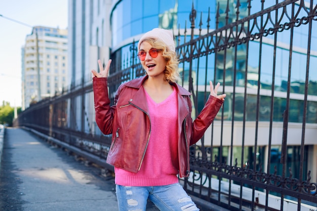 Outdoor portrait of joyful hipster enthusiastic woman in trendy pink hat, leather jacket . Showing signs by hand.
