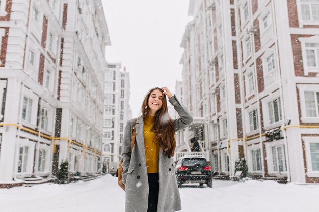 Outdoor portrait of long-haired woman in trendy gray coat going to shopping in snowy day. Gorgeous blonde woman in stylish outfit spending time in city in winter weekend.