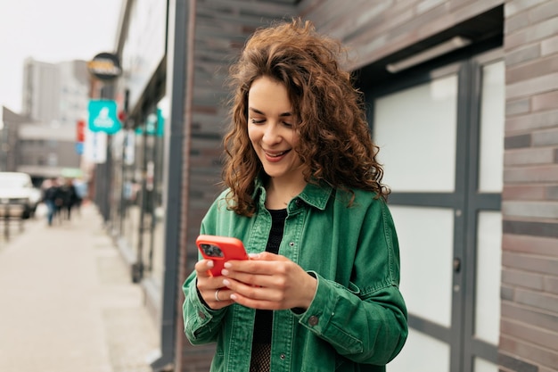 Открытый портрет прекрасной стильной девушки с кудрями в зеленой рубашке, использующей смартфон с улыбкой Беззаботная молодая кавказская девушка использует современный смартфон, стоящий на улице
