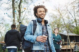 outdoor portrait of handsome unshaved dark-skinned guy standing in park with coffee, wearing headphones on neck and looking aside. man tries to find his girlfriend in crowd. emotions concept