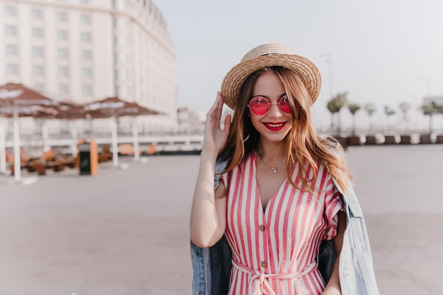Outdoor portrait of pleasant stylish girl in vintage hat standing on city background Goodhumoured female model in pink sunglasses enjoying warm spring day