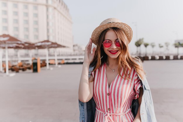 Открытый портрет приятной стильной девушки в винтажной шляпе, стоящей на фоне города Добродушная модель в розовых очках, наслаждающаяся теплым весенним днем