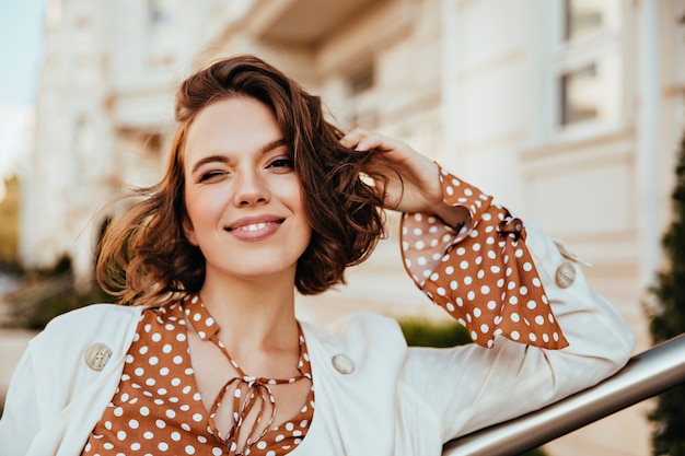 ぼやけた街で満足しているブルネットの女性の屋外の肖像画。トレンディなヘアカットで白人の魅惑的な女の子のショット。