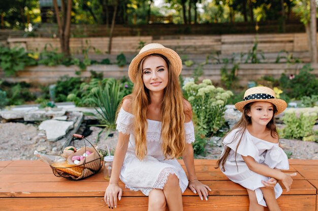 Outdoor portrait of pleased young woman and girl sitting with legs crossed in park on nature after picnic. Photo of charming lady with basket of food spending time with daughter in garden.