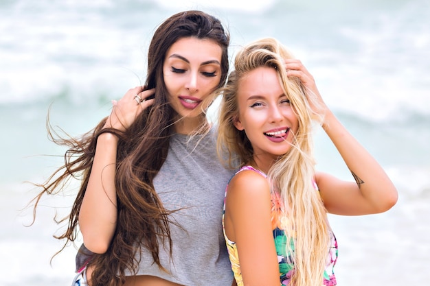 Outdoor portrait of pretty girl with dark-brown hair posing with eyes closed on blur sea background. Attractive blonde young woman with arm tattoo having fun with friend during summer vacation.