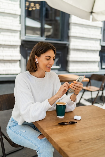 Outdoor portrait of pretty short haired woman enjoing cappucino in cafe, wearing cozy white sweater, listening to favorite music by earphones and chatting by mobile phone.