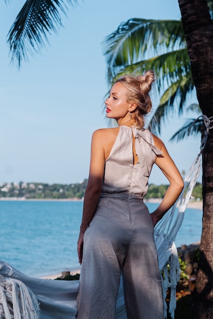 Outdoor portrait of rich caucasian woman in classic jumpsuit with red lipstick by hammock on vacation outside villa hotel