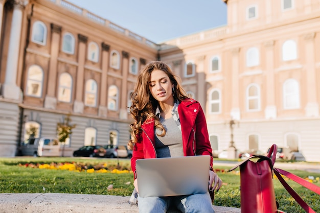 Outdoor portrait of serious curly female student sitting with laptop on the ground