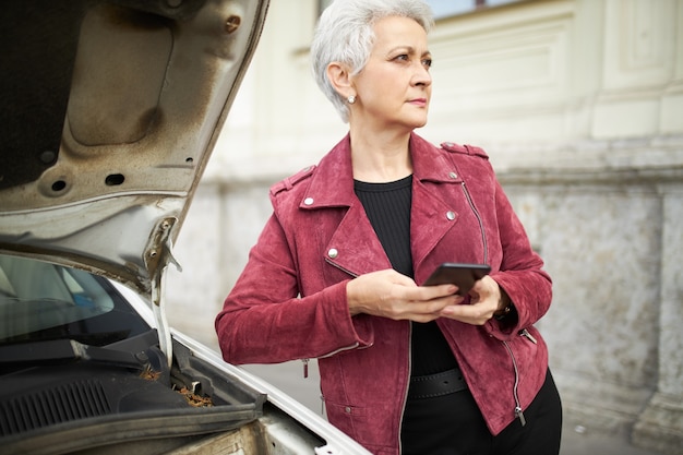 開いたフード、携帯電話を保持している彼女の壊れた車でポーズをとってスタイリッシュな服を着た真面目な成熟した実業家の屋外の肖像画