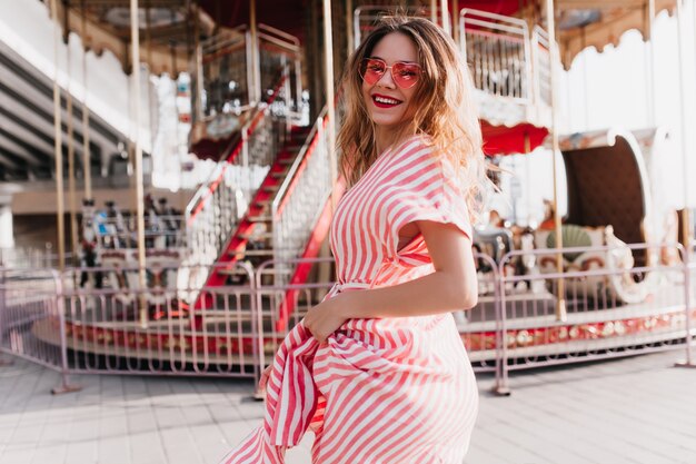 Outdoor portrait of shapely caucasian girl in summer attire posing in amusement park. Pleased female model in pink sunglasses standing near carousel and looking over her shoulder.