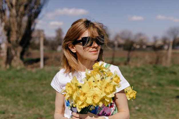 Il ritratto all'aperto di una ragazza sorridente in occhiali da sole tiene in mano fiori gialli distoglie lo sguardo e sorride donna alla moda in occhiali da sole che cammina all'esterno