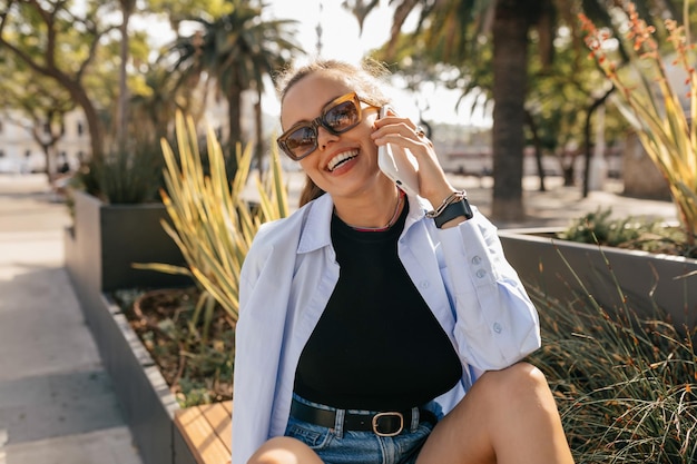 Открытый портрет улыбающейся счастливой девушки в синей рубашке и шортах, сидящей на солнечной улице со смартфоном и наслаждающейся солнечным днем