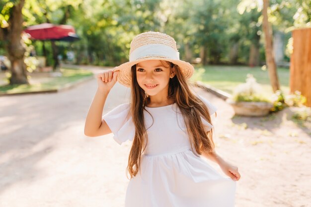 Outdoor portrait of smiling little girl with long straight dark hair walking in park in sunny morning. Cheerful female kid in straw hat and white dress enjoying vacation spending time on the street.