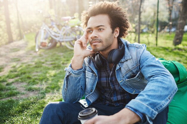 Outdoor portrait of stylish african man sitting in park with cup of coffee, holding hand on face and looking aside while thinking or dreaming