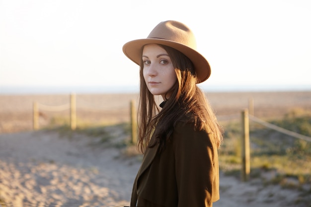 Ritratto all'aperto di elegante giovane donna europea che indossa un cappello alla moda e un cappotto nero guardando con un sorriso sottile mentre si fa una bella serata a piedi sul mare, sognando e ammirando il tramonto