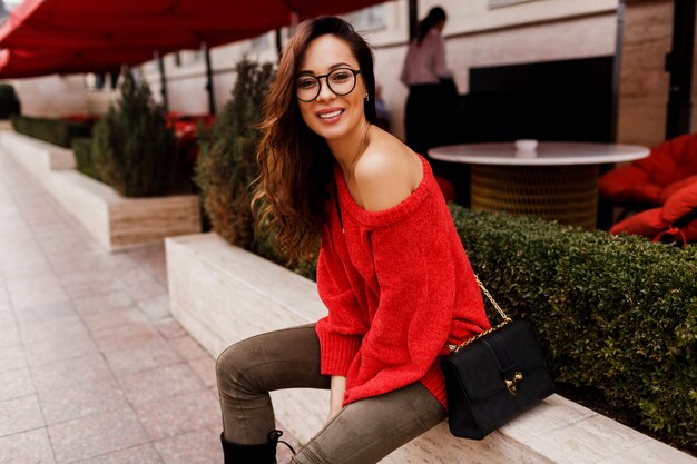 앉아 유럽 휴일을 즐기는 유행 빨간 니트 스웨터에 성공적인 웃는 갈색 머리 여자의 야외 초상화. 우아한 검은 색 가방.