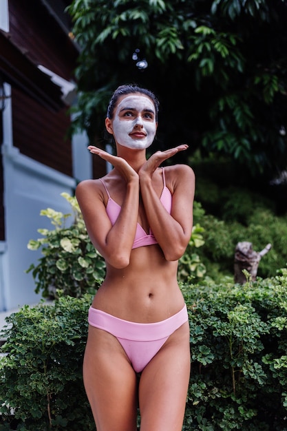 Outdoor portrait of tanned skin calm pretty caucasian woman in bikini at spa with white peeling mask on face