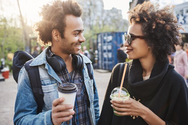 公園の周りにぶら下がって、コーヒーを飲みながら、笑って話している2人のかわいいアフリカ系アメリカ人の屋外のポートレート。