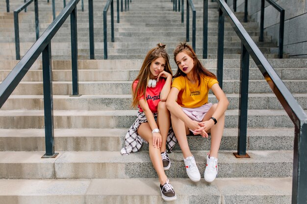 Outdoor portrait of two friends in stylish sneakers and bright shirts sitting on stairs