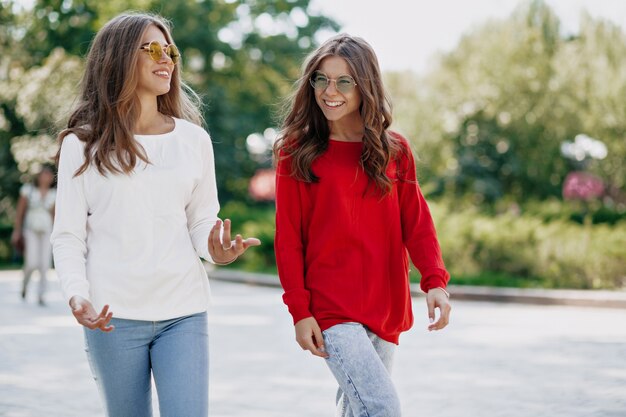 Outdoor portrait of two stylish sisters walking after shopping. Smiling long-haired girl in bright sunglasses talking and have fun in the city