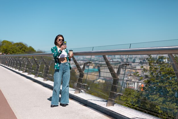 晴れた日のカジュアルな緑のシャツとジーンズの女性の屋外の肖像画は、電話の画面で見ているコーヒーと橋の上を歩く