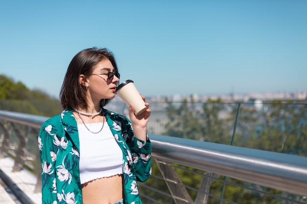 Outdoor portrait of woman in green shirt with cup of coffee enjoying sun, stands on bridge with city amazing view in morning