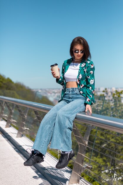 Outdoor portrait of woman in green shirt with cup of coffee enjoying sun, stands on bridge with city amazing view in morning