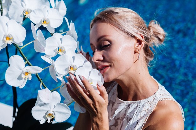 Outdoor portrait of woman in white wedding dress sitting near blue swimming pool with flowers orchid