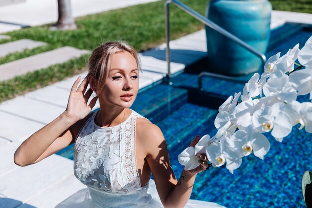 Outdoor portrait of woman in white wedding dress sitting near blue swimming pool with flowers orchid