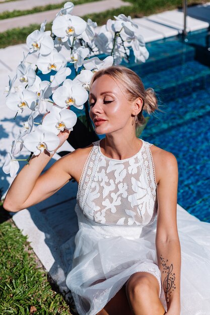 Outdoor portrait of woman in white wedding dress sitting near blue swimming pool with flowers